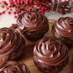 close up of wooden tray of finished chocolate cupcake with chocolate icing. Red berries in the background.