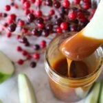 An apple slice dipped into homemade caramel sauce in a mason jar. Other apple slices in the background.