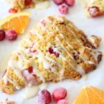 A close up of a finished cranberry orange scone with orange glaze. A parchment paper background and garnishes of cranberries and orange slices surround the finished scones.
