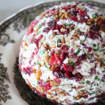 Close up of a finished cranberry pecan cheese ball in a vintage bowl, a lit candle in the background
