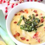 A white bowl of homemade potato leek soup, topped with bacon and fried leeks, with potatoes and leeks in the background.