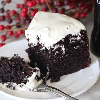 A finished slice of guinness chocolate cake with cream cheese frosting on a white plate. A forkful has been removed from the cake and it sitting on the plate in front. A bottle of Guinness Draught in the background.