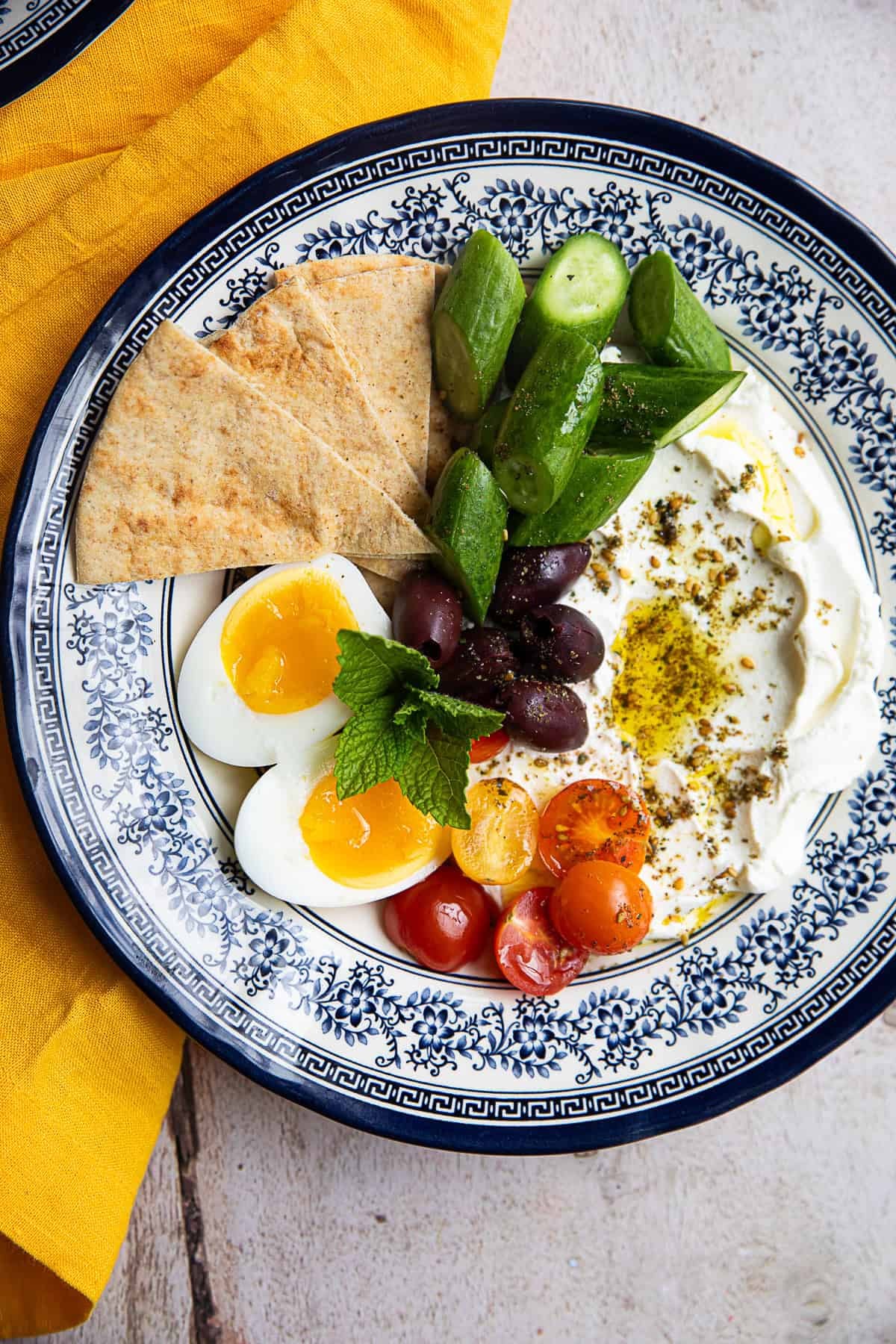 top down view of a blue and white plate with soft boiled eggs, pickles, olives, pita bread, and hummus
