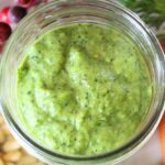 close up of a small mason jar with the finished carrot top pesto inside. Pine nuts, carrot greens, and carrots surround the jar