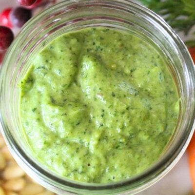 close up of a small mason jar with the finished carrot top pesto inside. Pine nuts, carrot greens, and carrots surround the jar