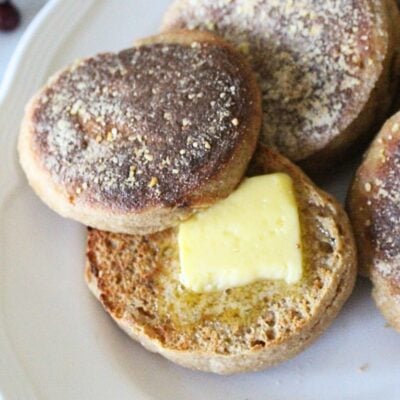 Finished homemade english muffins on a plate, one is cut open and toasted with melting butter on the middle.