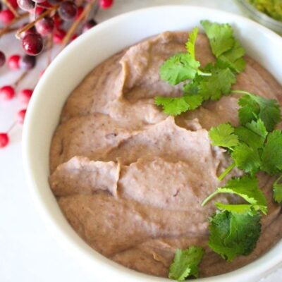 a white bowl of homemade refried beans, topped with cilantro garnish. Red berries in the background.