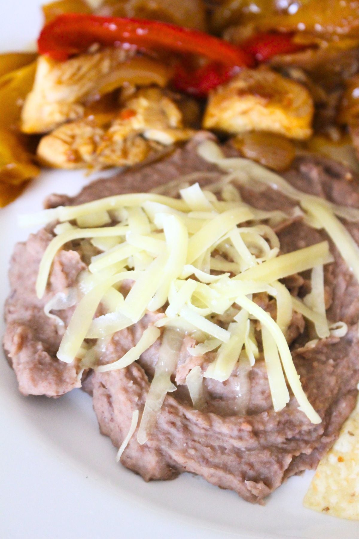 Finished refried beans on a plate, topped with cheese. Chicken fajitas in the background.