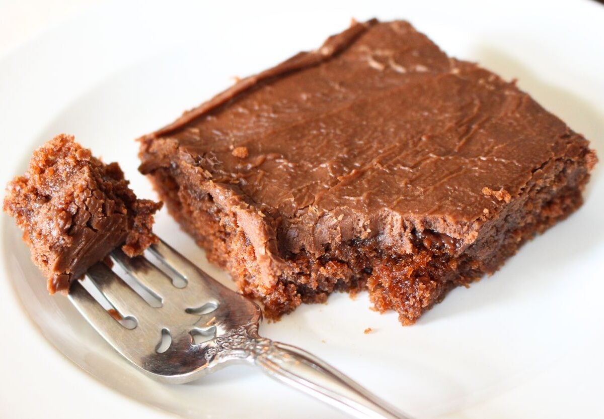 Finished slice of texas sheet cake on a white plate, a forkful has been removed and the fork with the bite are sitting beside the slice.