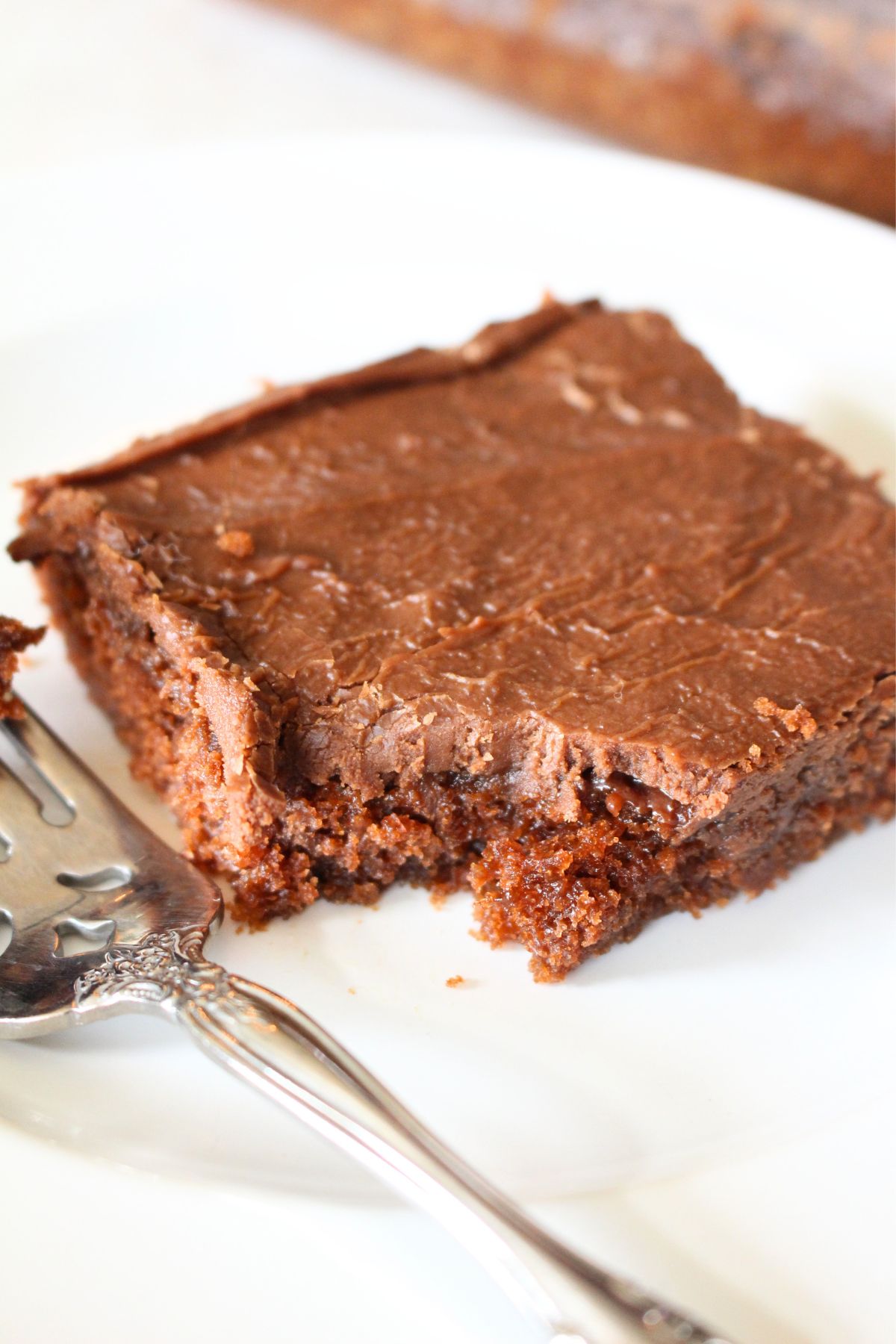 a square slice of texas sheet cake on a white plate, a bit missing from the edge and a fork beside the slice.