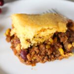 Finished serving of cornbread casserole, cowboy casserole in a white plate with the baking dish remainder in the background.